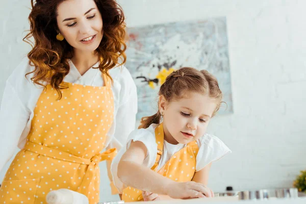 Glückliche Mutter in gelber Tupfen-Schürze, die Tochter in der Küche mit Teigformen betrachtet — Stockfoto
