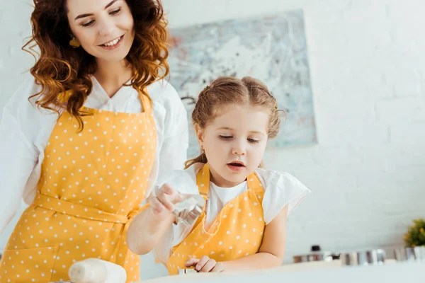 Mãe feliz em amarelo polka ponto avental olhando para a filha usando moldes de massa — Fotografia de Stock