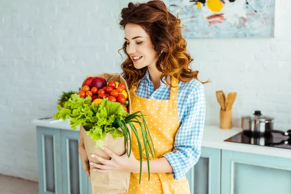 Donna sorridente in grembiule a pois con sacchetto di carta con frutta e verdura mature — Foto stock