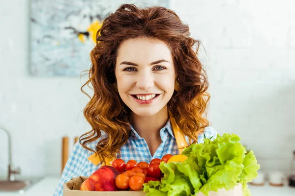 Retrato de una mujer sonriente cerca de una bolsa de papel con frutas y verduras maduras en la cocina - foto de stock