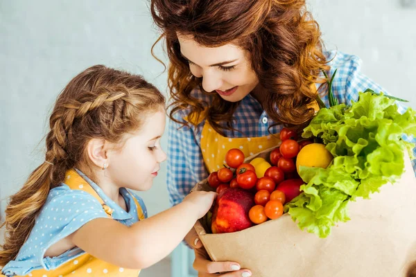 Madre feliz mostrando bolsa de papel hija con frutas y verduras maduras - foto de stock