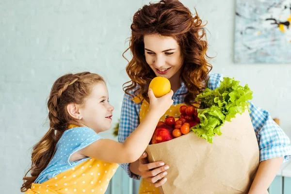 Smiling daughter giving lemon to mother with paper bag full of ripe fruits and vegetables — Stock Photo