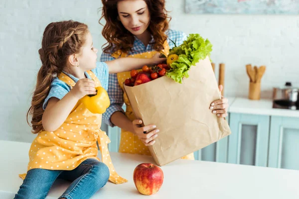 Hija tomando manzana y pimiento de la bolsa de papel en la cocina - foto de stock