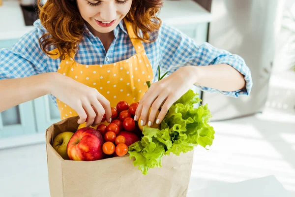 Femme heureuse à pois tablier jaune prenant des fruits et légumes dans un sac en papier — Photo de stock