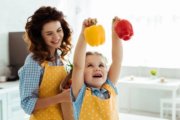 Mère heureuse regardant fille excitée tenant poivrons rouges et jaunes — Photo de stock