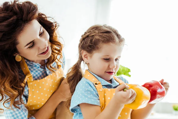 Mère heureuse regardant sa fille tenant des poivrons rouges et jaunes — Photo de stock