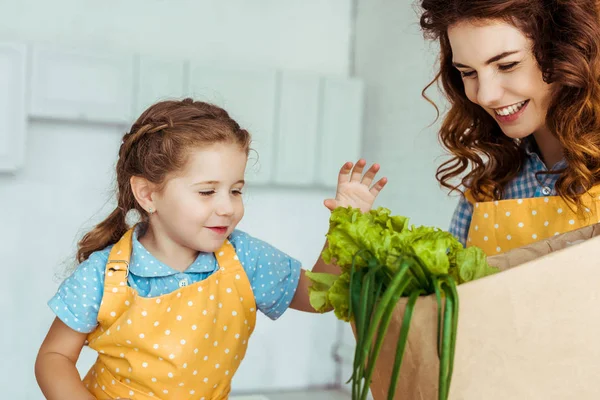 Glückliche Mutter in gepunkteter Schürze hält Papiertüte mit Salat neben lächelnder Tochter — Stockfoto