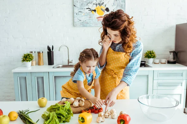 Madre e figlia in grembiuli a pois in piedi vicino al tavolo con frutta e verdura cruda — Foto stock