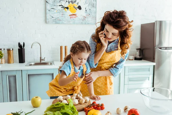 Madre hablando en el teléfono inteligente y mirando a la hija de pie cerca de la mesa con verduras - foto de stock