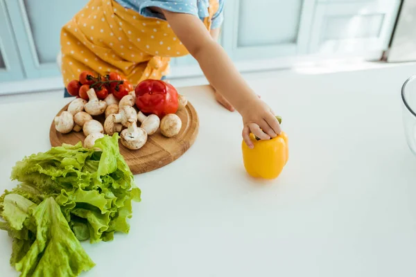 Cropped view of child in polka dot apron holding yellow bell pepper on table with vegetables and chopping board — Stock Photo