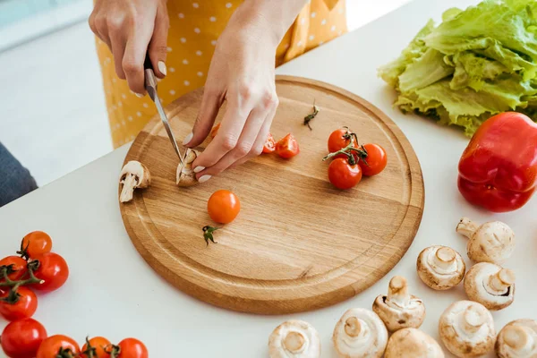 Cropped view of woman in polka dot apron cutting mushrooms and cherry tomatoes on chopping board — Stock Photo