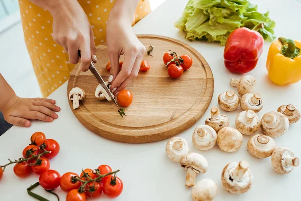 Vue recadrée de femme en tablier à pois coupant champignons et tomates cerises sur planche à découper près de la fille — Photo de stock