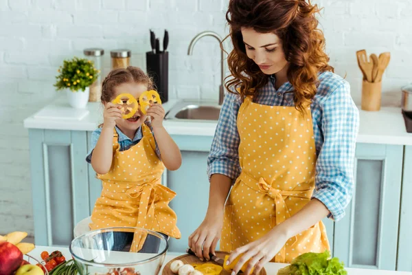 Linda hija sosteniendo rodajas de pimiento frente a los ojos mientras la madre corte verduras — Stock Photo