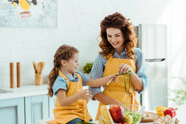 Linda hija en lunares delantal amarillo dando cebollas verdes madre - foto de stock