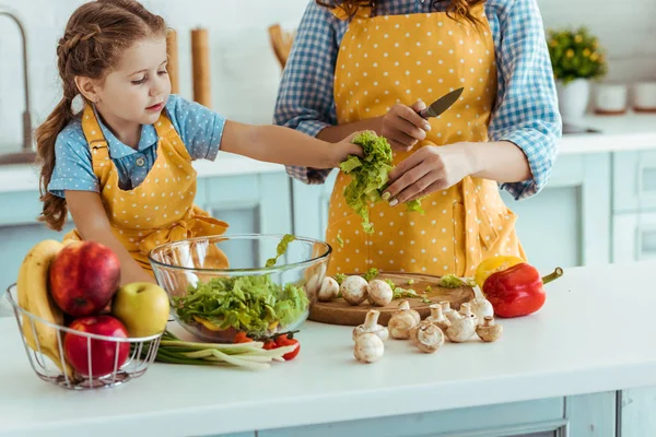 Mother in polka dot yellow apron giving daughter sliced lettuce — Stock Photo