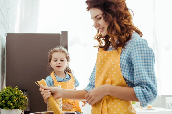 Neugierige Tochter blickt Mutter in gepunkteter gelber Schürze an und steckt rohe Spaghetti in Topf — Stockfoto