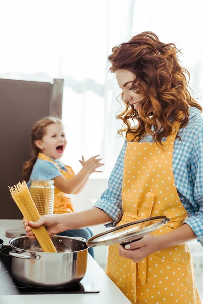 Enfoque selectivo de la madre en lunares delantal amarillo poner espaguetis crudos en olla mientras hija emocionada riendo de fondo - foto de stock