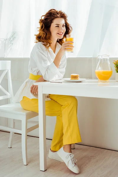 Belle femme heureuse assise à table et buvant du jus d'orange tout en prenant le petit déjeuner — Photo de stock