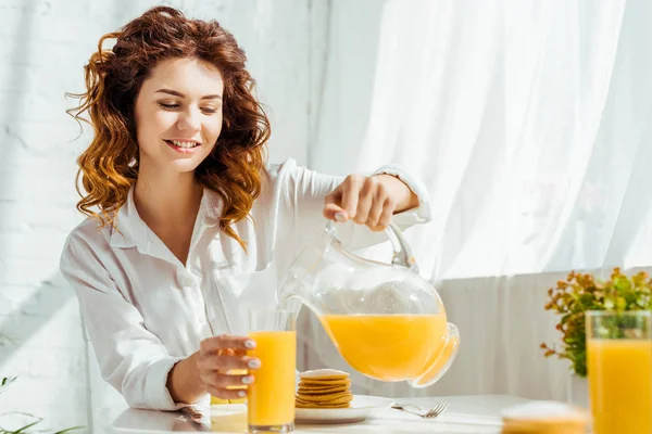 Hermosa mujer feliz sentada en la mesa y verter jugo de naranja en el vaso mientras desayuna - foto de stock