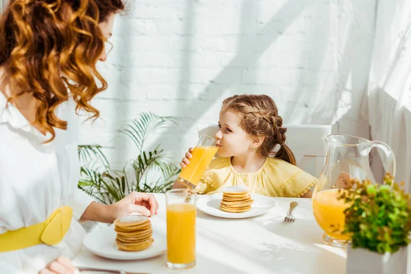 Lindo niño bebiendo jugo de naranja mientras desayuna con la madre - foto de stock