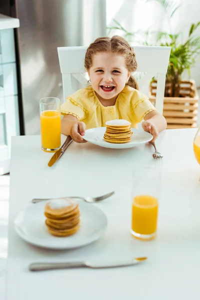 Selektiver Fokus des fröhlichen aufgeregten Kindes, das am Tisch neben Orangensaft sitzt und Teller mit Pfannkuchen hält — Stockfoto
