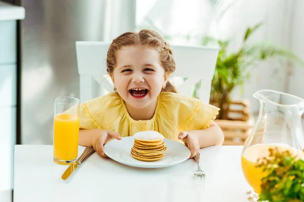 Happy excited kid sitting at table, laughing and holding plate with pancakes — Stock Photo