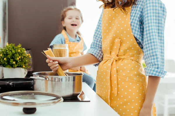 Vue recadrée de la femme à pois tablier jaune mettant spaghetti cru en pot avec fille excitée sur fond — Photo de stock