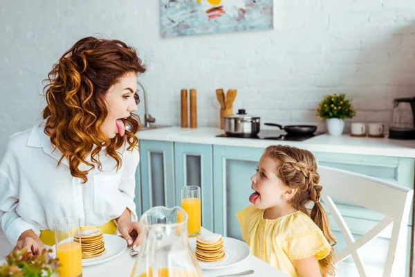 Drôle fille et mère sortant langues tout en prenant le petit déjeuner — Photo de stock