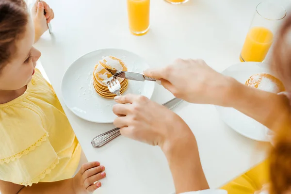 Cropped view of mother cutting pancakes with fork and knife near daughter — Stock Photo