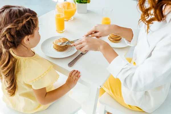 Teilansicht der Mutter beim Zuschneiden von Pfannkuchen mit Gabel und Messer in der Nähe der Tochter — Stockfoto