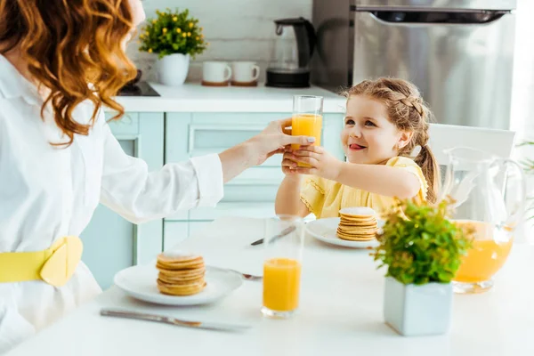 Enfoque selectivo de la madre dando vaso con jugo de naranja a la hija sonriente mientras desayuna - foto de stock
