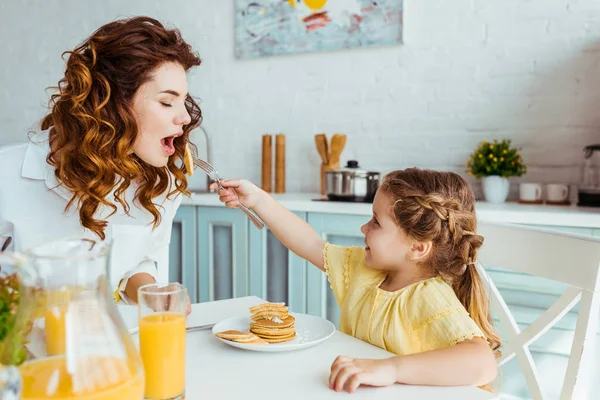 Smiling daughter feeding mother with pancakes during breakfast — Stock Photo