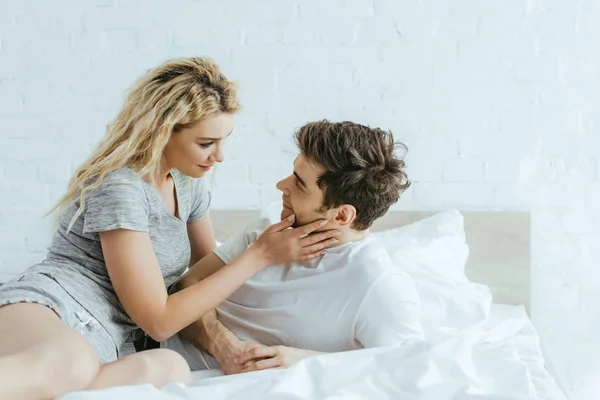 Attractive blonde woman touching cheek of handsome man lying on bed — Stock Photo