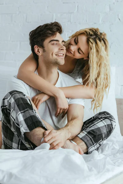 Attractive blonde woman hugging happy boyfriend sitting on bed — Stock Photo