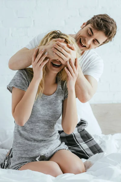 Hombre alegre cerrando los ojos de la mujer rubia feliz sentado en la cama - foto de stock