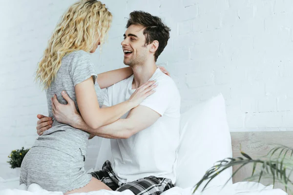 Happy handsome man looking at blonde girlfriend sitting on bed — Stock Photo