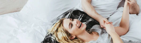 Panoramic shot of happy blonde woman holding hands with boyfriend while lying on bed — Stock Photo