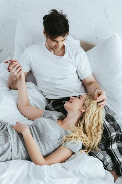 Glückliche blonde Frau hält Händchen mit hübschem Freund, während sie auf dem Bett liegt — Stockfoto