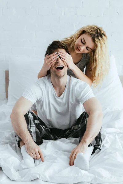 Attraktive blonde Mädchen schließt Augen eines glücklichen Mannes, der auf dem Bett sitzt — Stockfoto