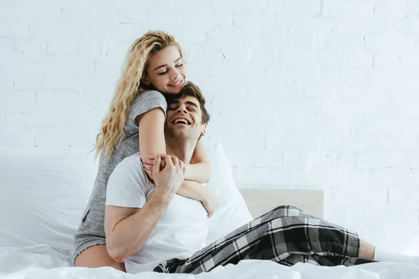 Привлекательная и счастливая блондинка обнимающая весёлого парня сидящего на кровати — стоковое фото