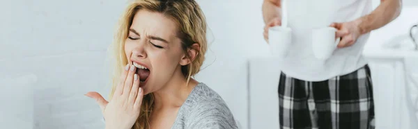 Panoramic shot of blonde woman yawning near man holding cups with drinks — Stock Photo