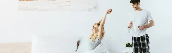 Panoramic shot of cheerful man holding cups near blonde woman stretching in bed — Stock Photo