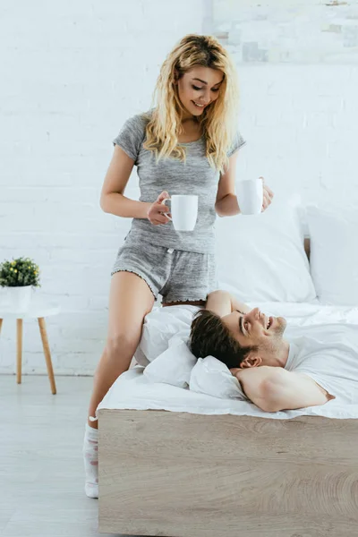 Alegre rubia mujer sosteniendo tazas con café cerca feliz hombre acostado en la cama - foto de stock