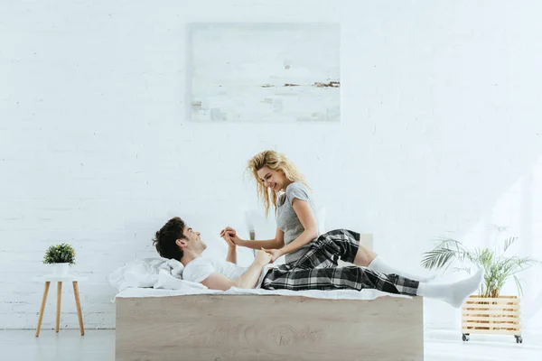 Happy blonde woman smiling while holding hands with man lying on bed — Stock Photo