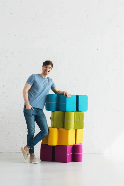 Cheerful man standing and smiling near colorful poufs near brick wall — Stock Photo
