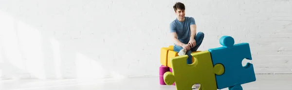 Plano panorámico de hombre alegre mirando piezas de rompecabezas azules y verdes mientras está sentado cerca de la pared de ladrillo - foto de stock