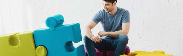 Panoramic shot of happy man looking at blue and green jigsaw pieces while sitting near brick wall — Stock Photo