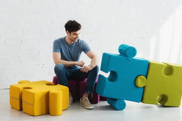 Cheerful man looking at connected blue and green jigsaw pieces while sitting near brick wall — Stock Photo