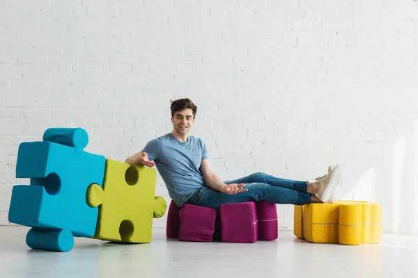 Cheerful man looking at camera near connected blue and green jigsaw pieces while sitting near brick wall — Stock Photo