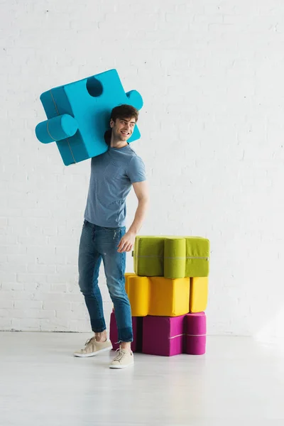Cheerful man standing and holding blue jigsaw piece near white brick wall — Stock Photo